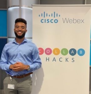 Charlie smiles in front of a sign advertising a Cisco Hackathon.