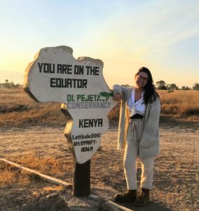 Cayla smiles next to a sign in the shape of Africa that says 'You are on the Equator' in Kenya.