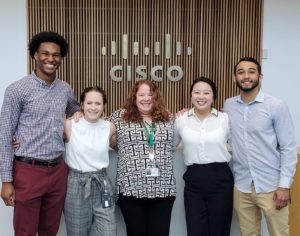 Emily smiles with her peers, a group of US Commercial new hires, in front of a Cisco lobby sign.