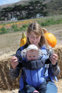 Carlene holds her baby in a carrier on her chest smiling in a pumpkin patch.