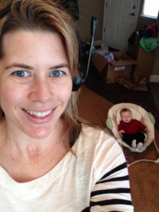 Carlene sitting at her desk at home with a headset on while her baby lays in a bassinet near her.