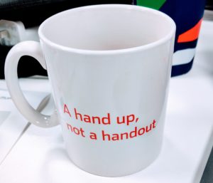 A white coffee mug with The Big Issue slogan 'A hand up, not a handout' in red on the side.