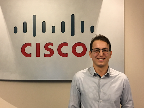 Samy smiles in front of a Cisco sign in glasses and a dress shirt.