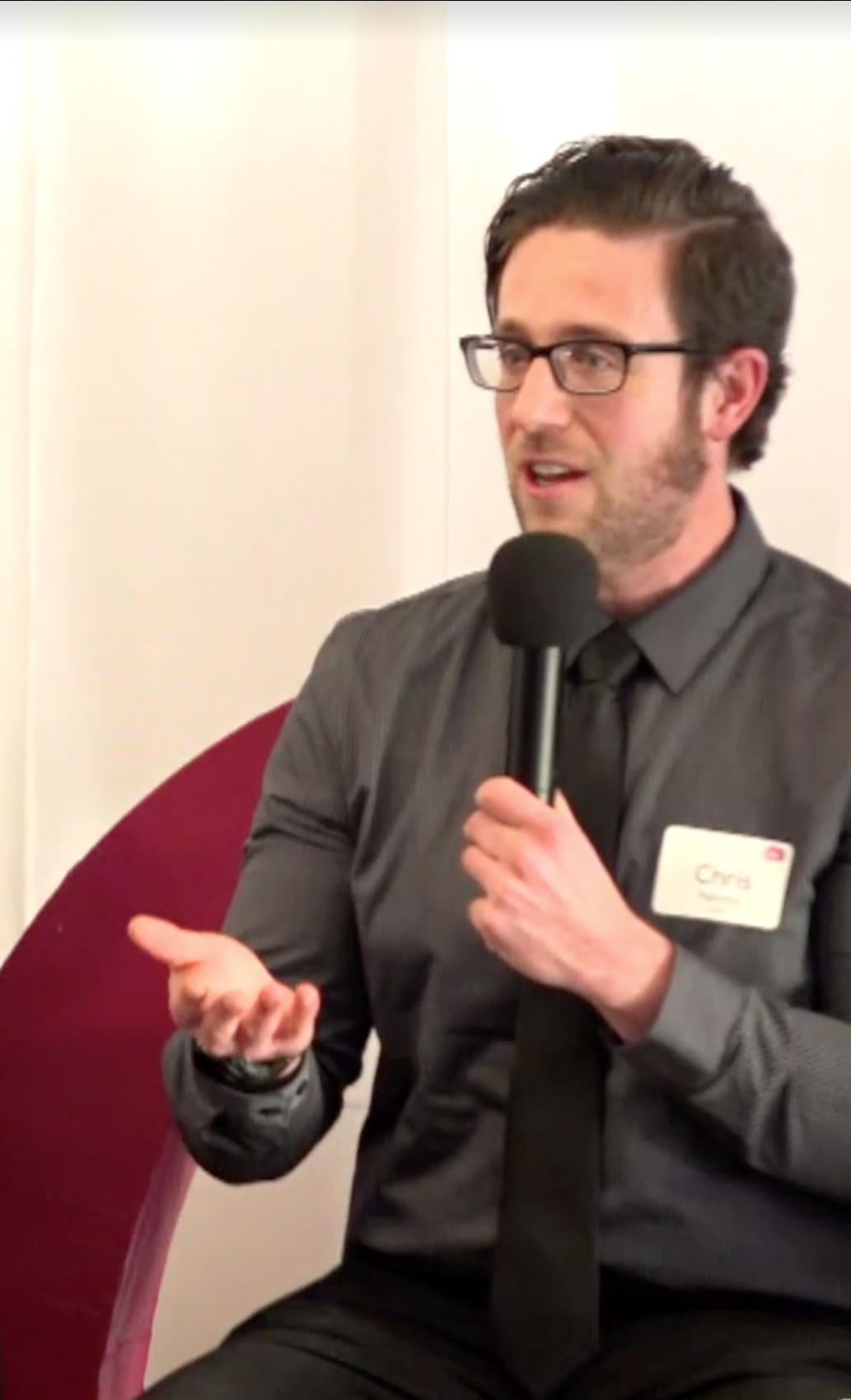 Chris holds a microphone while speaking in a shirt and tie.