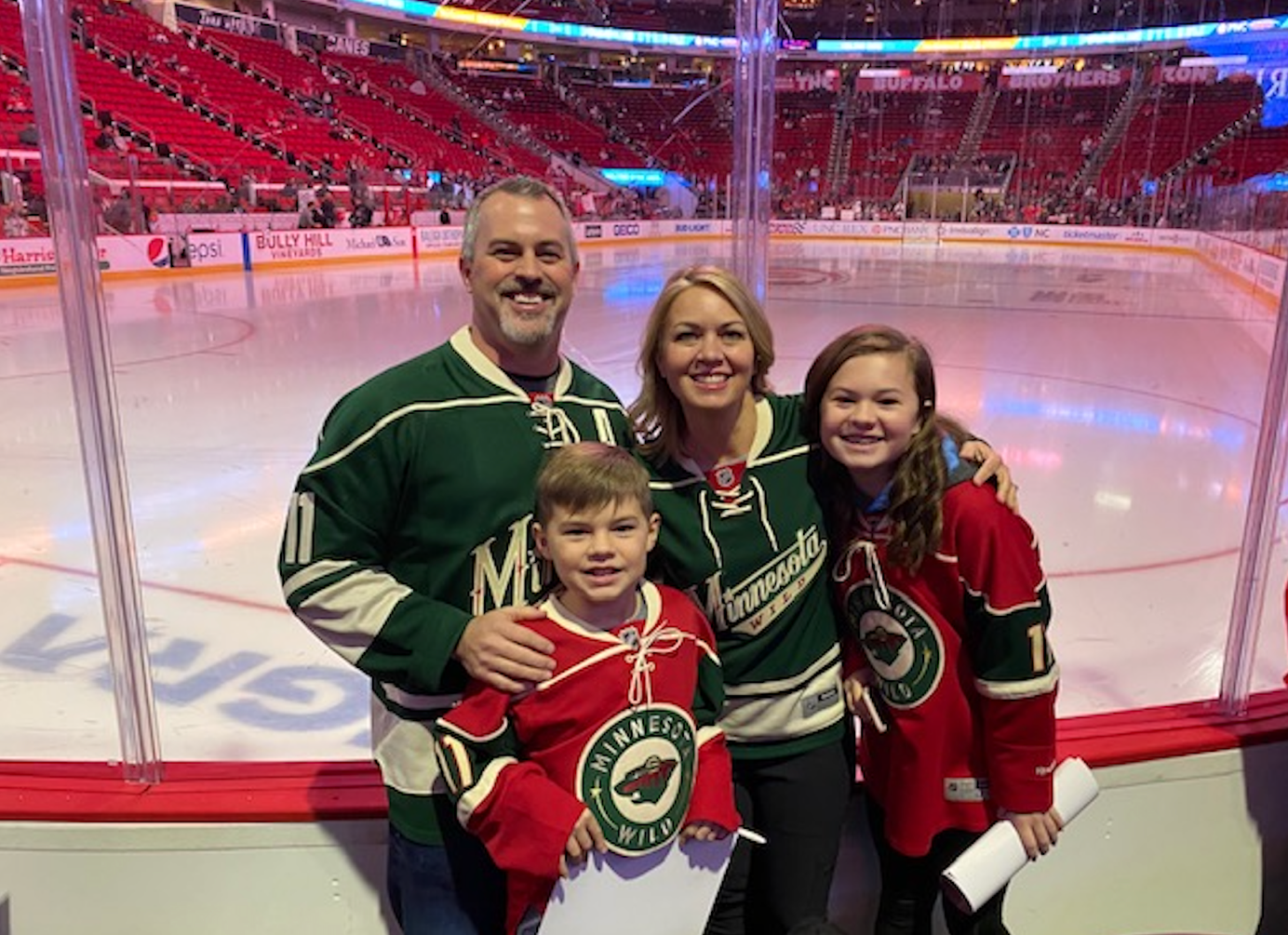 Shelly and her family pose next to the NHL ice hockey rink while all wearing Minnesota hockey jerseys.