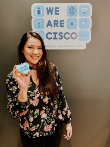 Ileana stands showing one of her sugar cookies that has blue icing and a Cisco logo on it.