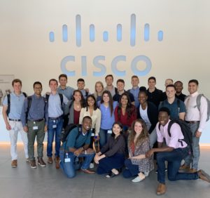 A group of Cisco interns take a photo in front of a Cisco logo.