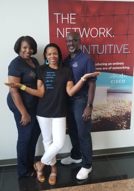 Shawnita, her husband Vanis, and her Mom Lovey all smile in their Cisco shirts in front of a Cisco advertisement. 
