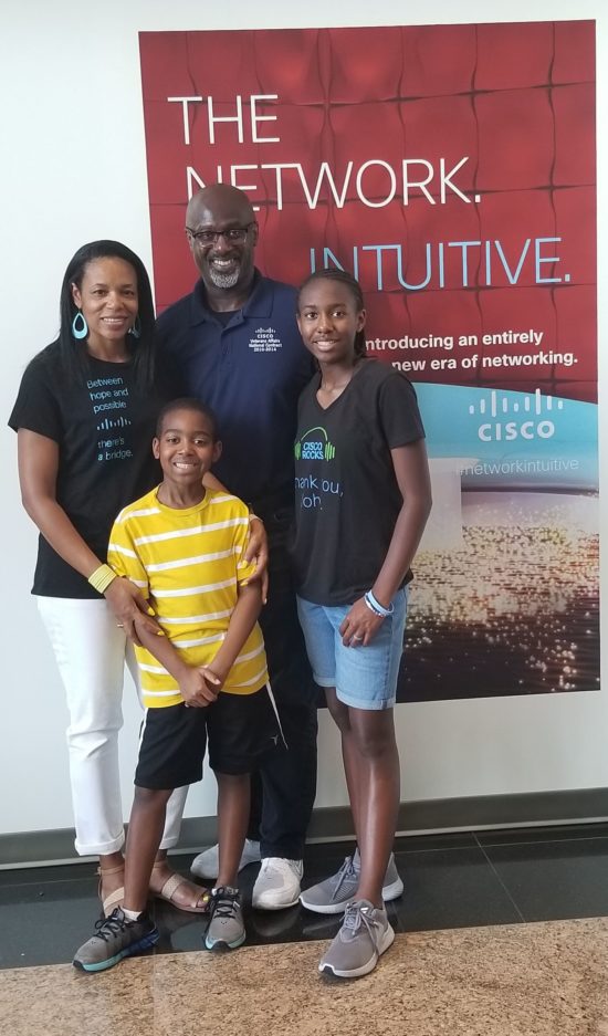 Shawnita stands with her husband, son, and daughter wearing Cisco shirts in front of a Cisco sign.