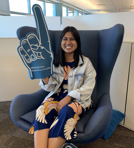 Alisha holds up a giant foam hand with the pointer finger raised and the hashtags #WeAreCisco and #LoveWhereYouWork.