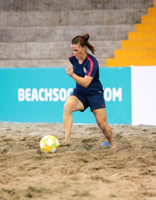 Jeane running after a soccer ball on a beach in her uniform.